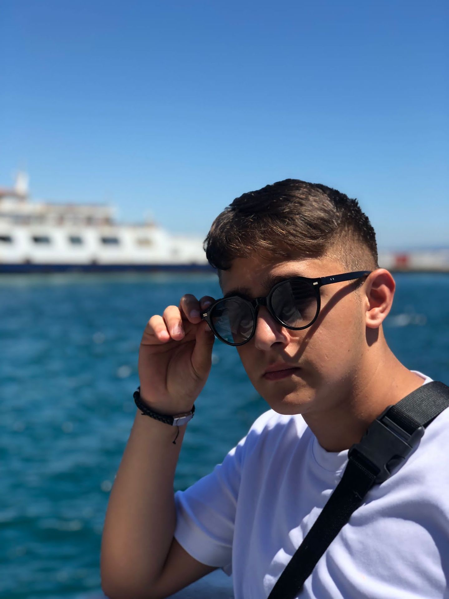 Person standing by the sea, holding sunglasses near their face, with a ferry in the background.