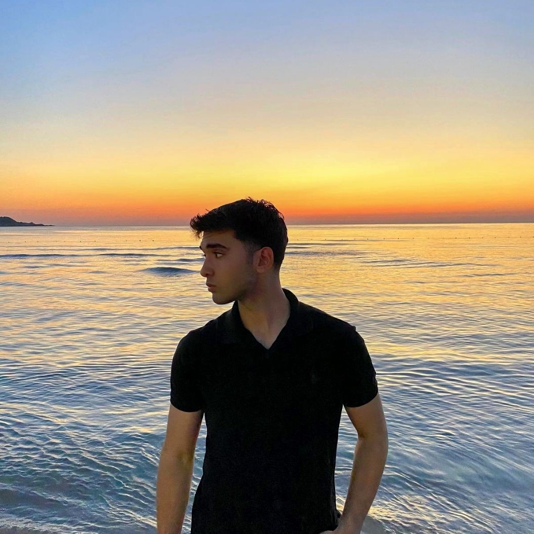 Person standing on the beach during sunset with calm sea and colorful sky in the background.